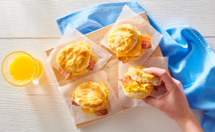 A hand reaching for one of four freshly made breakfast biscuit sandwiches filled with scrambled eggs, cheese, and bacon, placed on a wooden board with parchment paper. A blue cloth and a glass of orange juice are in the background.