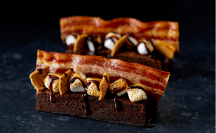 Two decadent brownies topped with crispy bacon strips, toasted marshmallows, and drizzled with chocolate, set against a dark background.