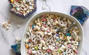 A white bowl filled with a snack mix of Lucky Charms cereal, yogurt-covered pretzels, popcorn, and colorful candy-coated chocolate pieces. The bowl is placed on a marble surface with smaller portions of the mix in decorative bowls nearby.