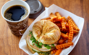 A croissant sandwich filled with turkey, cheese, and fresh greens, served with a side of crinkle-cut sweet potato fries in a white paper tray. A cup of black coffee in a disposable cup with a lid is placed next to the tray on a wooden table.