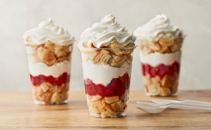 Three parfait cups filled with layers of crispy cereal, creamy yogurt, and strawberry compote, topped with a generous swirl of whipped cream. A white plastic spoon lies next to the cups on a wooden surface.